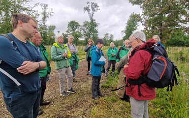 A group gathers around a man in red, talking amongst a grassy area of new woodland.