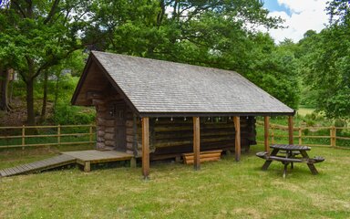 A log cabin in a grassed area surrounded by a wooden fence with trees behind