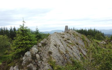 A trig point on top of a rocky outcrop above a forest