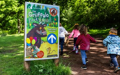 Four children with their backs to the camera walking down a forest path next to a promotional sign for Gruffalo Party Trail
