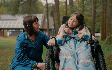 A young girl in a wheelchair in the forest, with her mother crouched next to her.