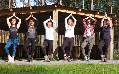 A group of six people standing in 'tree' yoga pose in a forest