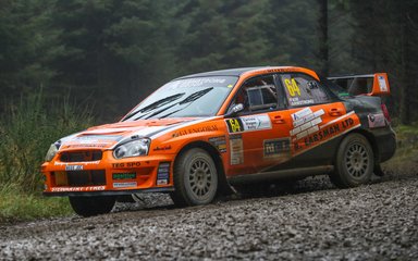 An orange rally car on a gravel track with trees in the background