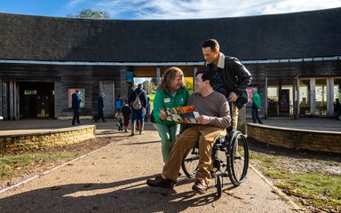 Two men, one standing and one in a wheelchair speaking to a lady in green in front of building whilst looking at a leaflet 