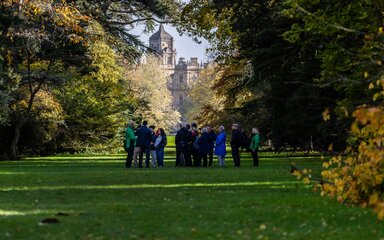 A group stand around listening to a guided walk in the distance. Behind them in the far distance is a grand building.