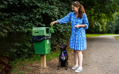  A woman in a blue dress putting a dog poo bag in poo bin watched on by her black dog 