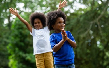 2 young brothers stand one in front of the other. The older brother in a white top lifts his arms in the air while the younger brother in a bright blue top grins with his hands together