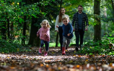 A young boy and girl run towards the camera over crunchy brown leaves with their parents smiling in the background.