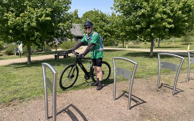 A picture showing a man in green on a black bike with bike racks in foreground and grass and trees behind