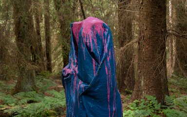 A mixed media sculpture that looks like a cloth stood up in the forest covered in blue and pink paint.