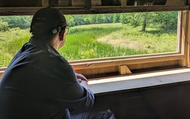 A man looking out of a wide glass-less window over a green meadow area surrounded by trees