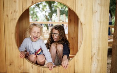 A young girl and boy look cheery through a wooden circular window.