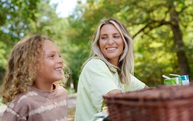 A woman and young children smiling over a wicker picnic hamper, with trees in the background.