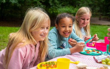 Three young girls with plates of fruit on a gingham-covered table outside.