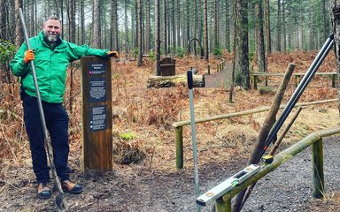 A white man wearing a green outdoor jacket and dark trousers leaning on a wooden post in the forest, with tools shown around him.