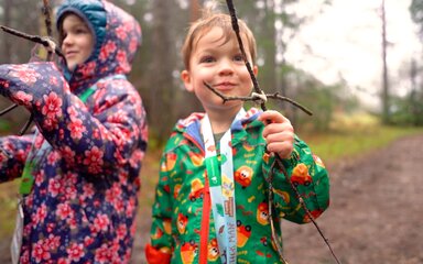 Two children on a forest path holding figures made from sticks.