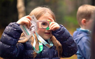 A girl in the forest looking through a viewfinder attached to a lanyard she's wearing.