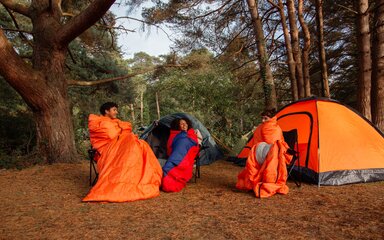 Three people sit in orange sleeping bags outside on orange tent in the forest.