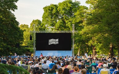 A big screen sits in front of trees, with a black image with the logo of Adventure Cinema. A crowd of people sit in front of the screen.