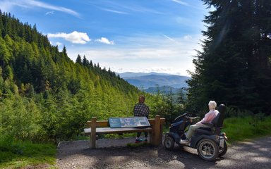 With their backs to the camera, a person sitting on a bench and a person sitting on an all-terrain mobility scooter look out over a high viewpoint