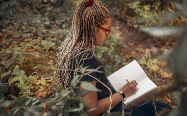 Side view of a women sat on a leafy forest floor, writing in a notebook