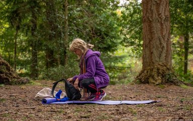 A woman crouches on a yoga mat looking at equipment in a forest clearing.