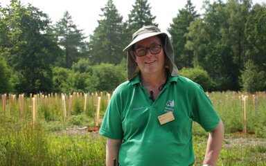 A male volunteer in a green polo shirt and sunhat stands smiling in the sun, surrounded by colourful tree tubes