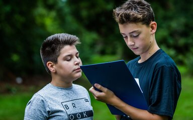 Two boys looking at a clipboard, surrounded by trees.