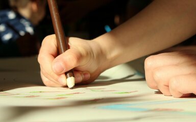 Close-up of a child's hand holding a coloured pencil, drawing on paper.