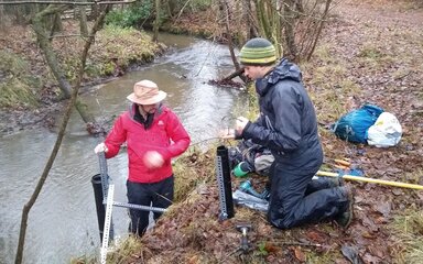 Two men with water monitoring equipment on a muddy river bank.