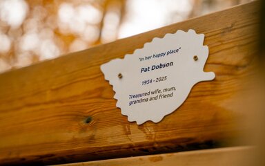 A silver-coloured memorial plaque in the shape of a leaf, in position on a wooden bench.