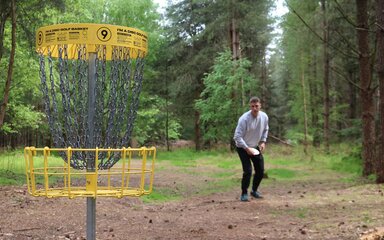A man holding a flying disc close to a disc golf net in the forest.