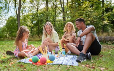 A family of two adults and two children in the forest, sitting with a picnic on a picnic rug. They are smiling at each other. 