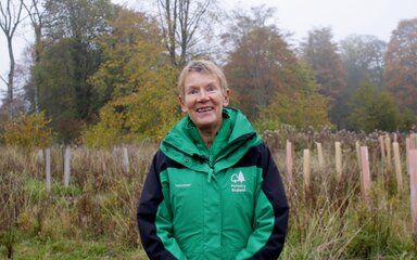 A female volunteer in a green jacket stands smiling surrounded by colourful tree tubes on a murky autumn day.