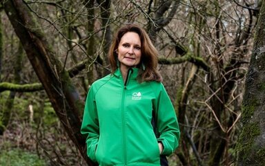 A white woman with long brown hair standing in woodland, wearing a green Forestry England jacket.