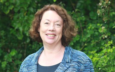 Upper torso and headshot of a smiling woman with short brown wavy hair, standing in front of green bushes.