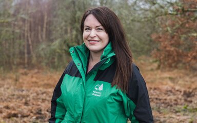 Headshot of a white woman with long dark hair, wearing a Forestry England branded coat, standing outdoors.