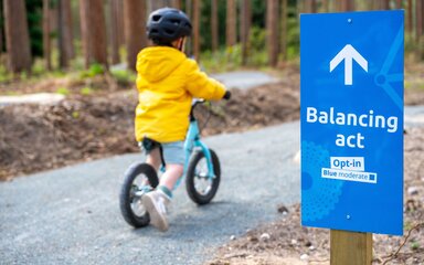 Young boy riding a blue balance bike along a forest track.