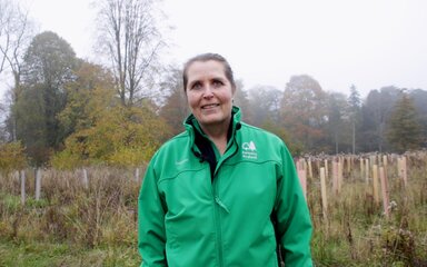 A female volunteer in a green jacket stands smiling surrounded by colourful tree tubes on a murky autumn day.