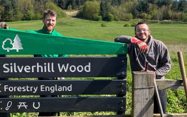 Two volunteers smiling as they learn on a wooden Forestry England sign saying Silverhill Wood