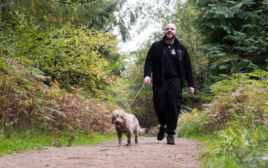 A man smiling as he walks a dog on a lead, on a forest path.