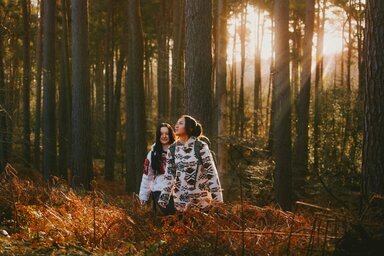 Two women in the forest at dusk, with low sun shining through tall tree trunks.