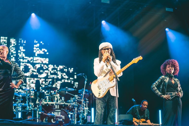 Male singer Nile Rodgers on stage, holding an electric guitar and singing into a microphone.