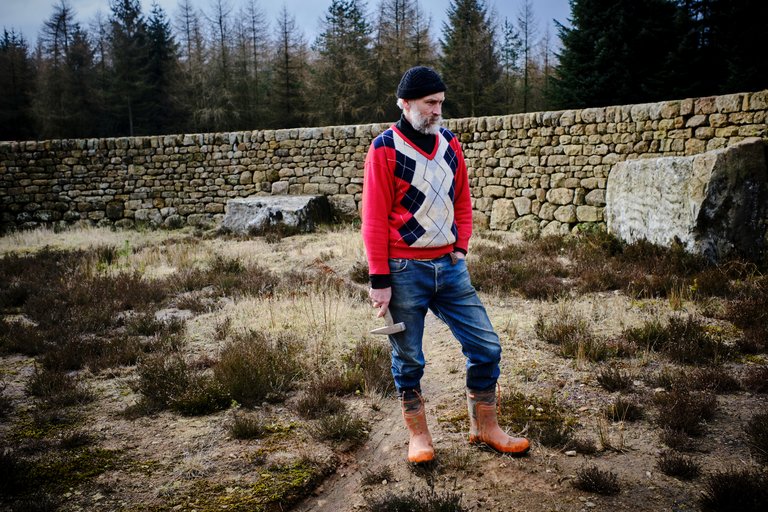 Man stood staring at the ground holding a wooden tool within stone wall maze